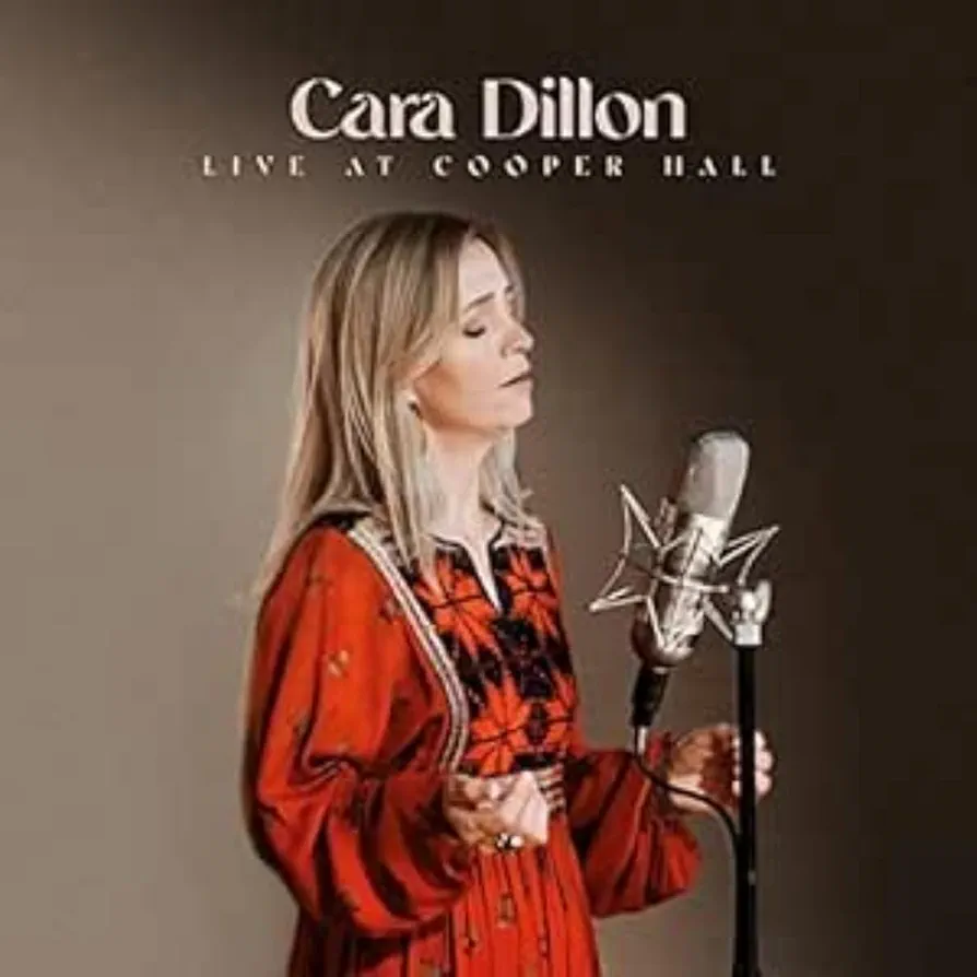 A woman stands in front of a microphone wearing a red dress, with text above her that reads "Cara Dillon Live at Cooper Hall."