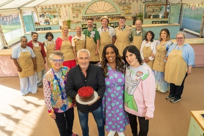 A group of people, including judges and contestants, stand together in a brightly decorated baking tent with one holding a cake.