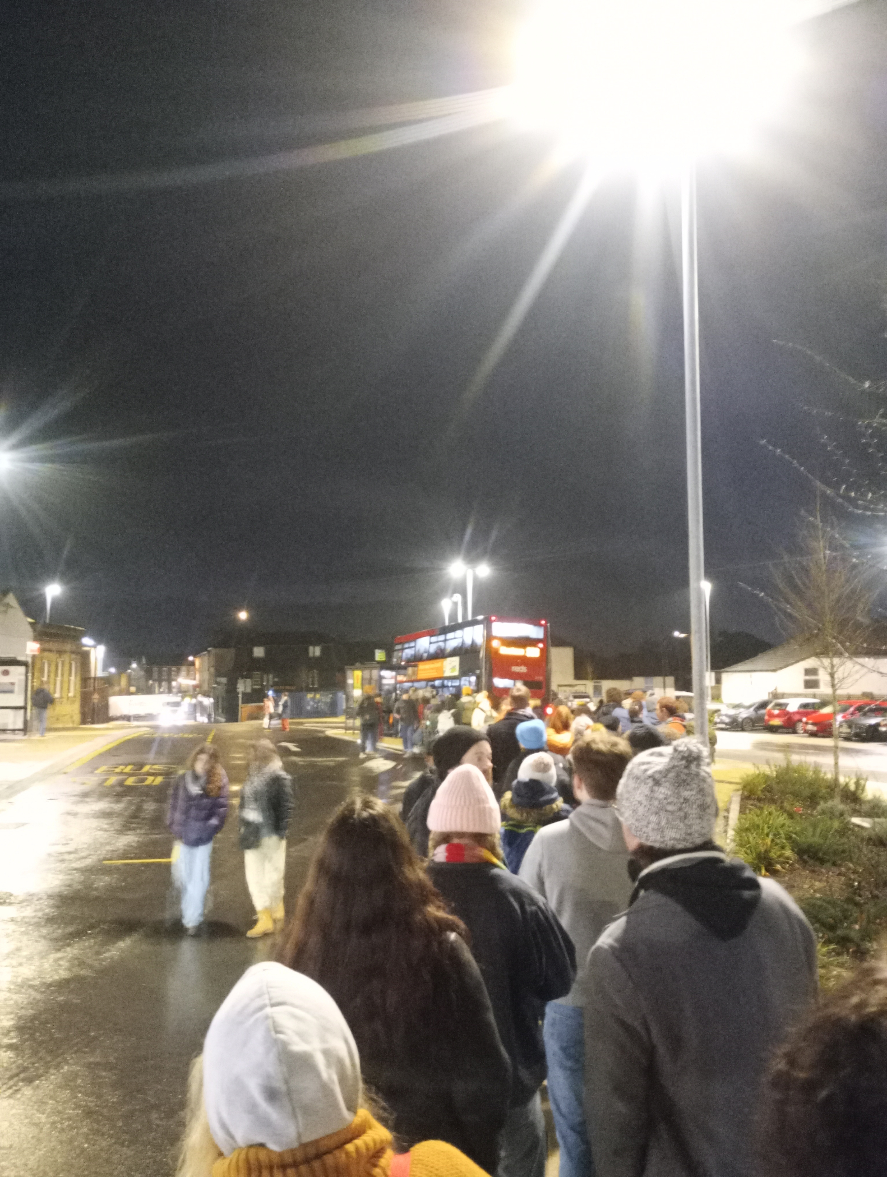 People are standing in a line at night, waiting for a bus at a well-lit stop.
