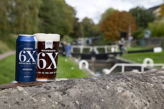 A can and a glass of Wadworth 6X Original Ale sit on a stone ledge by a canal with blurred background scenery of greenery and locks.